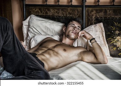 Shirtless sexy male model lying alone on his bed in his bedroom, looking away with a seductive attitude