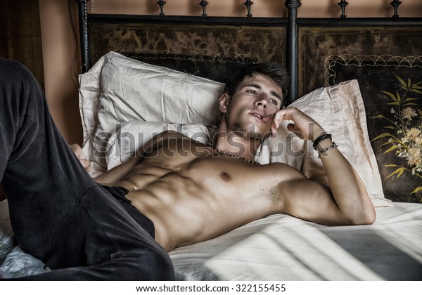 シャツのないセクシーな男性モデルが寝室のベッドに一人で寝そべり 誘惑的な態度で目をそらす の写真素材 今すぐ編集