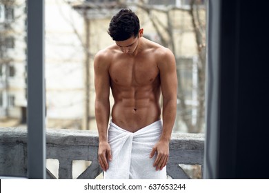 A shirtless sexy strong fit muscular young man in bathroom towel, showing six pack abs, standing on the balcony, isolated on street background. Horizontal view.
