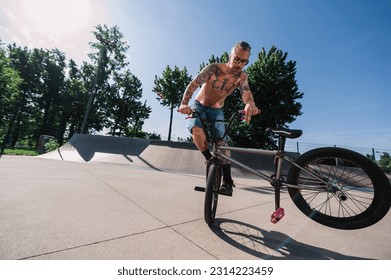 A shirtless tattooed mature man is performing tricks and stunts in a skate park with his bmx. A mature urban freestyle bike rider is practicing tricks and stunts on his bike in a skate park.