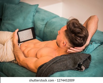 Shirtless young athletic man holding ebook reader or tablet PC and reading while sitting on couch - Powered by Shutterstock