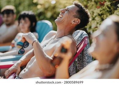 Shirtless young man laughs while reclining on a lounge chair holding sunglasses. Friends relax together outdoors on colorful chairs in a sunny summer setting.