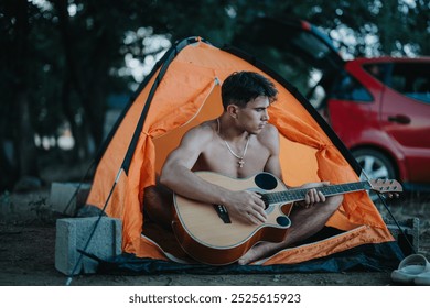 Shirtless young man playing guitar inside an orange tent while camping in nature. Relaxing and enjoying the outdoors. - Powered by Shutterstock