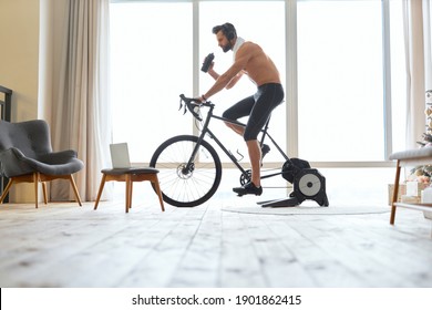 Shirtless young man riding stationery bike and drinking water