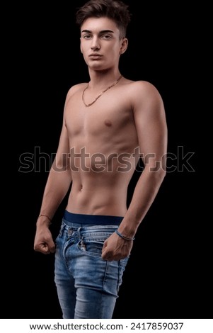 Shirtless young man with toned physique sports golden chain and confident gaze against a dark background