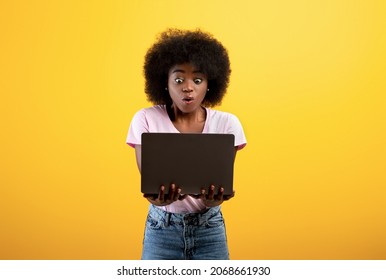 Shock Content. Overwhelmed Black Lady Looking At Laptop Screen, Holding Pc Computer, Receiving News Or Suffering From Internet Scam, Standing On Yellow Background