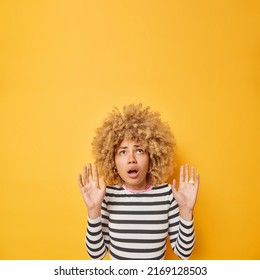 Shocked Adult European Woman Keeps Arms Up Like Something Falling From Above Focused On Ceiling Dressed In Casual Striped Jumper Isolated Over Yellow Background Empty Space For Your Promotion