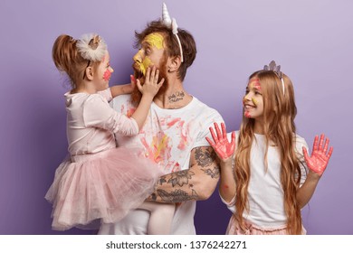 Shocked Affectionate Dad Has Dirty Face From Paints, Opens Mouth Widely, Holds Little Daughter On Hands. Smiling Girl Wears Crown, Shows Palms In Pink Watercolors. Joyful Father And Children Have Fun