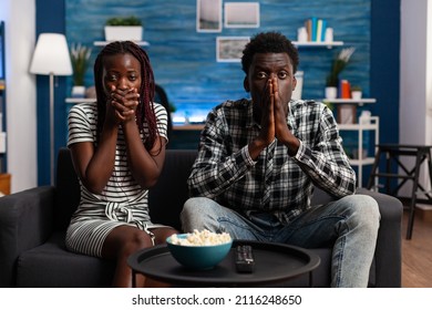 Shocked african american couple watching tragic news on television program while sitting at home. Terrified people reacting bad while watching horror movies sitting alone on couch.