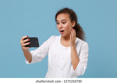 Shocked African American Doctor Woman Isolated On Blue Background. Doctor In Medical Gown Doing Selfie Shot On Mobile Phone Make Video Call. Healthcare Personnel Medicine Concept. Mock Up Copy Space