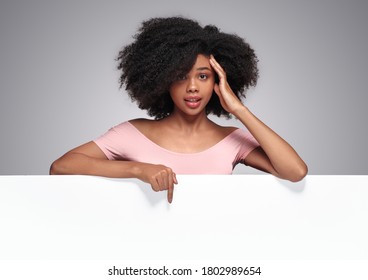 Shocked African American Female With Curly Hair Touching Head And Looking At Camera While Demonstrating Empty Banner During Skin Care Service Advertisement Campaign