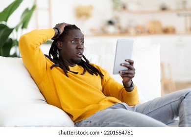 Shocked African American Guy Looking At Digital Tablet Screen At Home, Confused Young Black Man Emotionally Reacting To Bad News While Sitting With Modern Gadget On Couch In Living Room, Free Space