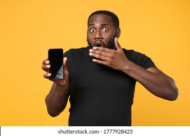 Shocked African American Man Football Fan In Casual Black T-shirt Isolated On Yellow Background Studio. People Lifestyle Concept. Hold Mobile Phone With Blank Empty Screen, Covering Mouth With Hand