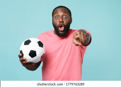 Shocked African American Man Football Fan In Pink T-shirt Isolated On Blue Background. Sport Family Leisure Concept. Cheer Up Support Favorite Team With Soccer Ball, Pointing Index Finger On Camera