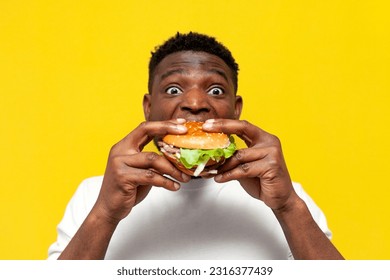 shocked african american man in white t-shirt holding big burger and surprised, amazed guy eating fast food on yellow isolated background, emotions from delicious food - Shutterstock ID 2316377439