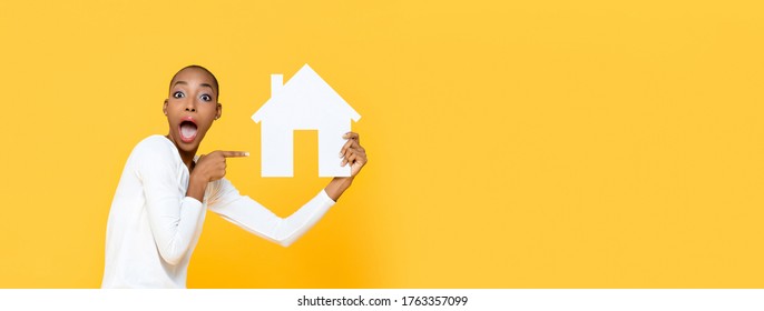 Shocked African American Woman Gasping And Pointing To House Cutout Model Isolated On Yellow Banner Background With Copy Space