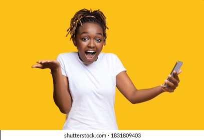 Shocked African American Young Lady With Smartphone On Yellow Studio Background, Won Lottery, Copy Space. Young Black Woman Gambling Online, Using Mobile Phone, Won The Prize, Celebrating Success