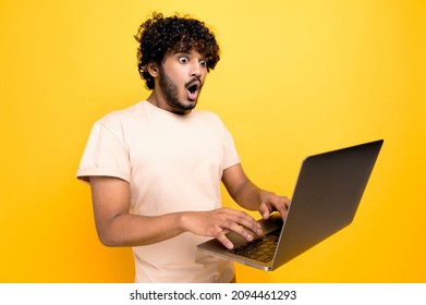 Shocked amazed indian or arabic curly-haired guy, typing on a laptop, looking in surprise at the screen, saw unexpected news, victory, profit, stands on isolated orange background. Emotional face