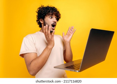 Shocked amazed indian or arabic curly-haired guy, looking in surprise at the laptop screen, saw unexpected news, victory, profit, standing on isolated orange background, gesturing hands.Emotional face