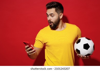 Shocked amazed surprised young bearded man football fan in yellow t-shirt cheer up support favorite team hold soccer ball use mobile cell phone isolated on plain dark red background studio portrait