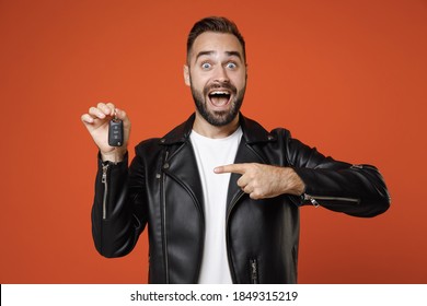 Shocked Amazed Young Bearded Man 20s In Basic White T-shirt, Black Leather Jacket Standing Pointing Index Finger On Car Keys Looking Camera Isolated On Bright Orange Colour Background Studio Portrait