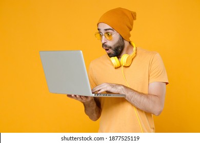Shocked Amazed Young Bearded Man 20s Wearing Basic Casual T-shirt Headphones Eyeglasses Hat Standing Hold Working On Laptop Pc Computer Isolated On Bright Yellow Colour Background, Studio Portrait