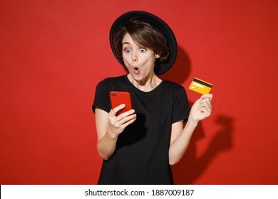 Shocked Amazed Young Brunette Woman 20s Years Old Wearing Casual Basic Black T-shirt Hat Standing Using Mobile Cell Phone Hold Credit Bank Card Isolated On Bright Red Color Background Studio Portrait