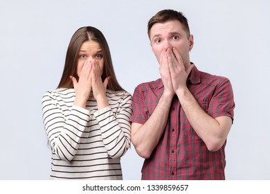 Shocked Amazed Young Couple Covering Mouth With Hands Looking At Camera. Stunned Surprised Man And Woman Astonished About Unexpected Information. Studio Shoot.