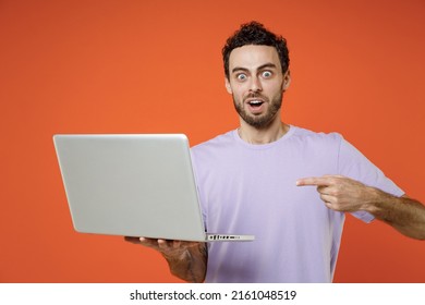 Shocked Amazed Young Man In Casual Basic Violet T-shirt Standing Working Pointing Index Finger On Laptop Pc Computer Look Camera Isolated On Orange Background Studio Portrait. Tattoo Translate Fun