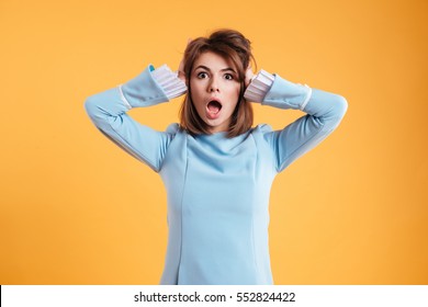 Shocked amazed young woman with hands on head standing and shouting over yellow background