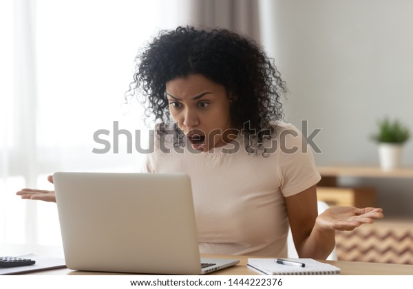 Shocked angry stressed african black woman customer\
looking at laptop screen feel bad surprise annoyed reading online\
news frustrated with stuck computer problem website error sit at\
home office desk