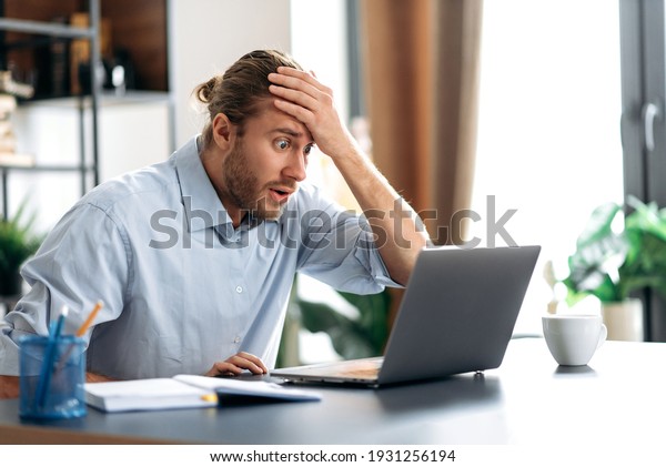Shocked annoyed\
caucasian busy guy looking at a laptop in confusion, received\
unexpected news, failed a deal, shouts, holds his head with his\
hand, sitting at the work\
desk