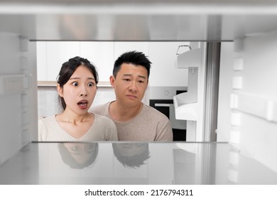 Shocked Asian Couple Looking Inside Open Empty Fridge In Kitchen At Home. Poor Chinese Family Husband And Wife Have Nothing To Eat, Copy Space. Financial Hangover While Crisis, Poverty Concept