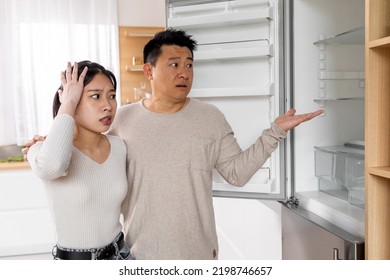 Shocked Asian Couple Standing Next To Open Empty Fridge In Kitchen At Home, Touching Head And Gesturing. Poor Chinese Family Husband And Wife Have Nothing To Eat, Copy Space