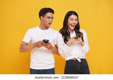Shocked asian man spying on her smiling girlfriend while both using mobile phones isolated over yellow background. chatting or social media concept. - Shutterstock ID 2128068167