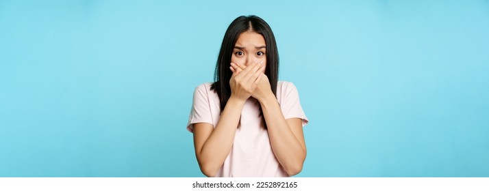 Shocked asian woman covers mouth screams, gasping startled, scared of smth, shuts her lips, standing over blue background. - Powered by Shutterstock
