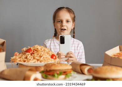 Shocked astonished little girl with braids sitting at table with junk food isolated over gray background looking at camera with big eyes uaing smartp phone online food delivery.