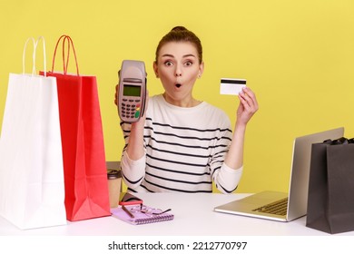 Shocked Astonished Woman Office Employee Sitting At Workplace With Shopping Bags, Showing Terminal And Credit Card To Camera. Indoor Studio Studio Shot Isolated On Yellow Background.