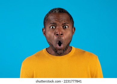Shocked attractive african american middle aged man with open mouth looking at camera, checking something astonishing on blue studio background, closeup portrait. Human emotions concept