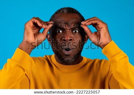 Shocked attractive african american middle aged man with open mouth removing eyeglasses, checking something astonishing on blue studio background, closeup portrait. Human emotions concept