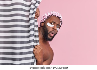 Shocked bearded dark skinned man hides behind shower curtain has amazed face expression applies beauty patches under eyes has daily hygiene procedures isolated over pink background. Cleanliness