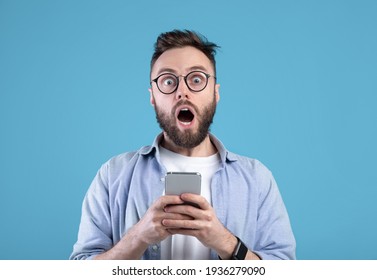 Shocked Bearded Guy Staring At Mobile Phone In Disbelief Over Blue Studio Background