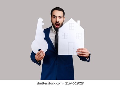 Shocked Bearded Man Holding Paper House And Big Key, Real Estate Purchase, Rental Services, Mortgage, Wearing Official Style Suit. Indoor Studio Shot Isolated On Gray Background.