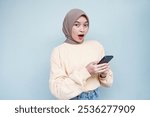 Shocked beautiful Asian muslim woman in cream sweater and hijab using a mobile phone, received good news isolated over white background. People religious lifestyle concept.