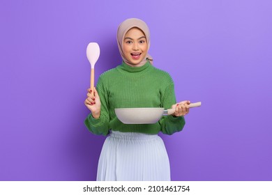 Shocked Beautiful Asian Woman In Green Sweater And Hijab Holding Spoon And Pan While Doing Housework Isolated Over Purple Background