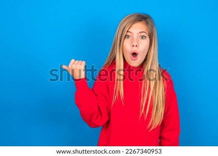 Shocked Beautiful caucasian teen girl wearing red sweatshirt over blue background points with thumb away, indicates something. Check this out. Advertisement concept.