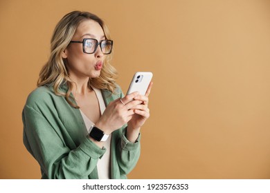 Shocked beautiful woman making kiss lips and using mobile phone isolated over beige wall