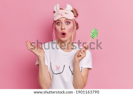 Shocked beautiful young European woman with blonde hair keeps mouth opened has bright vivid makeup wears sleepmask and casual t shirt holds lollipop isolated over pink background. Omg concept