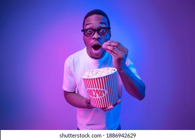 Shocked black guy in 3D glasses eating popcorn in cinema under neon light, funny surprised african american man watching interesting movie and looking at camera with excitement over studio background - Powered by Shutterstock