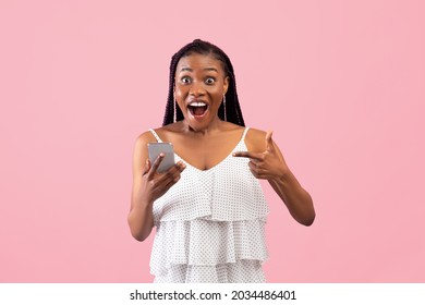 Shocked black lady pointing at smartphone in excitement, winning lottery or online casino bet on pink studio background. Excited African American woman receiving amazing news on phone